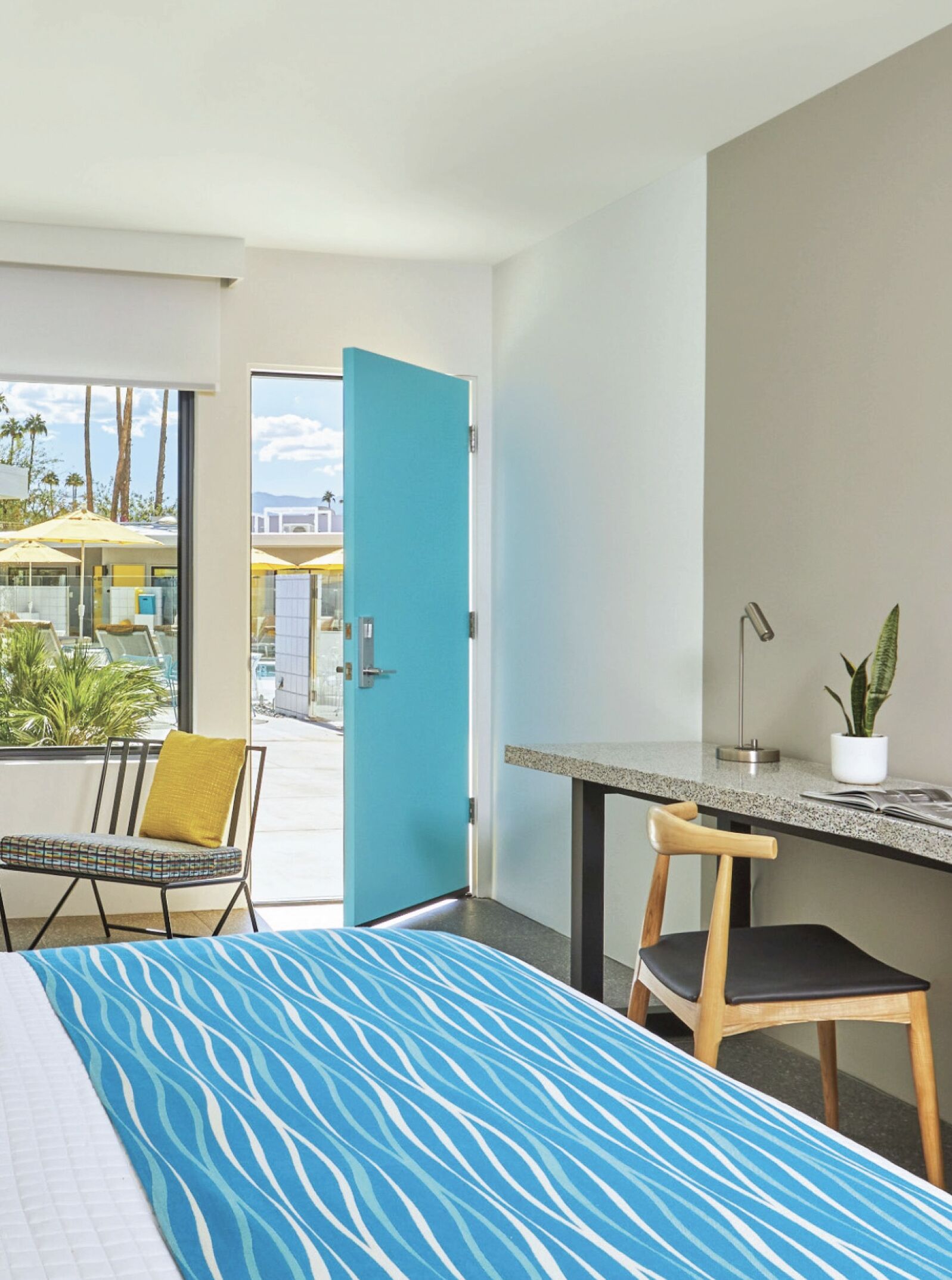 Camera d'hotel con un letto, una scrivania, una sedia, una pianta in vaso e vista sulla zona piscina.