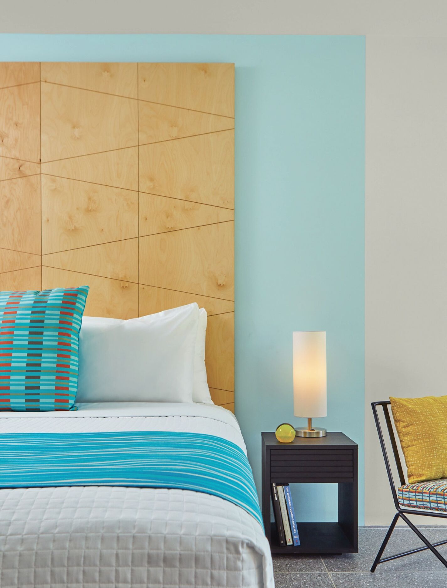 A hotel room with a wooden headboard, a bed with striped bedding, a nightstand with a lamp, and a chair with a yellow cushion.