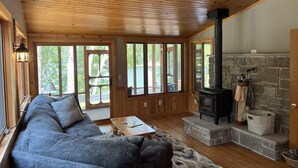 L’intérieur chaleureux d’un chalet avec un plafond en bois, une cheminée en pierre, un canapé et de grandes fenêtres donnant sur une forêt.
