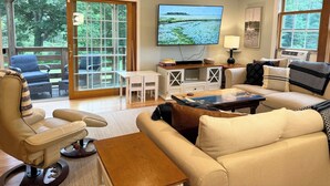 A cozy living room with a large TV, comfortable seating, and a view of greenery through the glass doors. - Family Lake Getaway on Davis Stream  5BR + Kayaks (Jefferson)