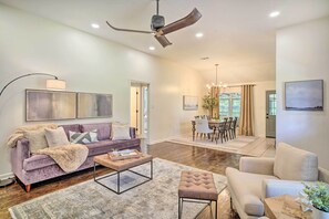 A spacious living room with a purple sofa, a beige armchair, and a coffee table. There is a dining area with a chandelier and a view of the outdoors. - Charming Baton Rouge Retreat ~ 3 Mi to LSU! (Baton Rouge)