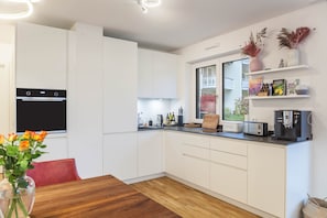 Une cuisine moderne avec des meubles blancs, un four encastré, une machine à café et une fenêtre donnant sur un balcon.