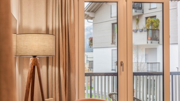 Una habitación con una lámpara de pie, una silla y un ventanal corredero que da a un balcón exterior con valla blanca y edificios al fondo.