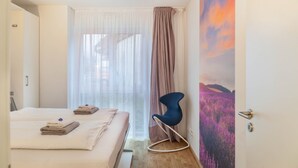 Habitación de hotel con dos camas, una silla, un ventanal con cortinas traslúcidas y un mural en la pared.