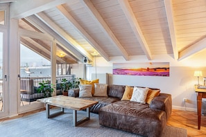 Un salon chaleureux avec un grand canapé en cuir marron, une table basse en bois et une vue sur un balcon extérieur, accessible par une porte vitrée.