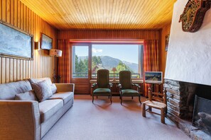 Une pièce chaleureuse avec un plafond en bois, une cheminée, un canapé, deux fauteuils et une grande fenêtre donnant sur les montagnes.