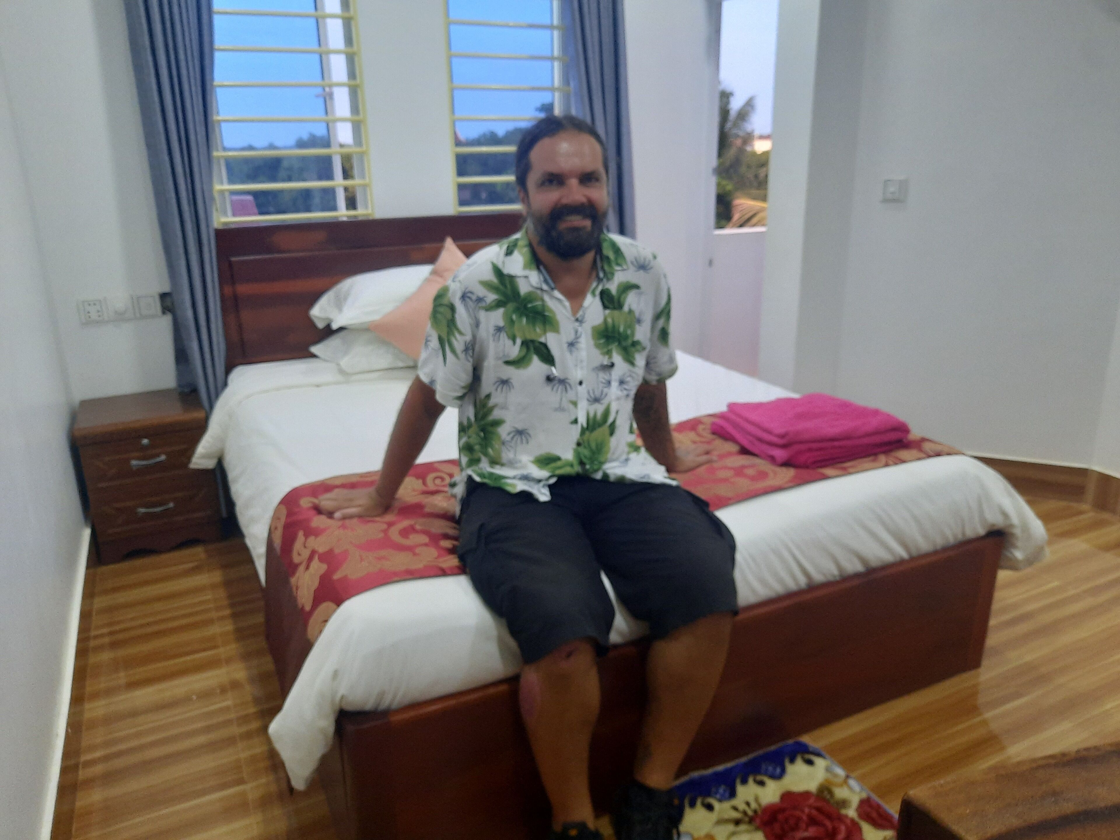 A man sitting on a bed in a hotel room with a wooden floor, a window with curtains, and a bedside table with a lamp.