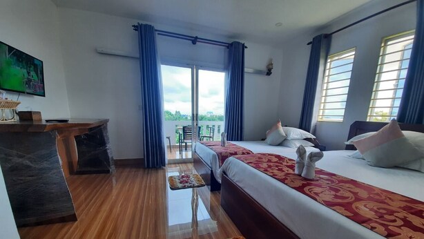 A hotel room with a wooden floor, a bed with a red and gold bedspread, a desk with a computer, and a view of the outdoors through large windows. - Realkhom Villa (Siem Reap)