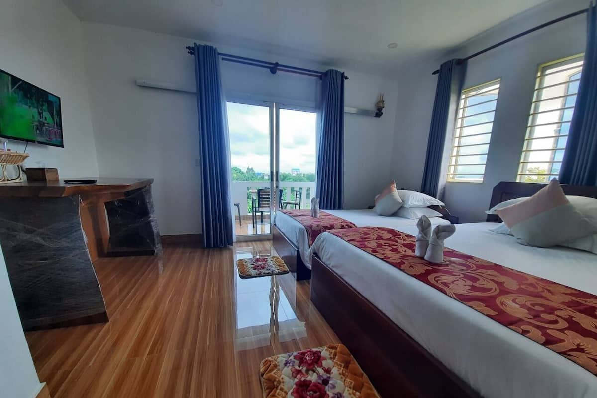 A hotel room with a wooden floor, two beds with red and white bedding, a small table with a floral runner, and a view of the outdoors through large windows.