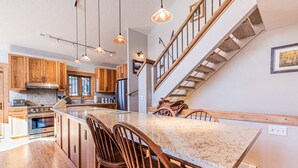 A kitchen with wooden cabinets, a large island with bar stools, a stainless steel refrigerator, and a staircase with a wooden railing.