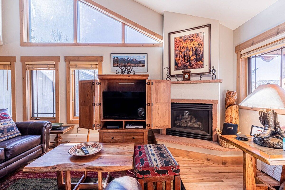 A living room with a fireplace, wooden furniture, a television, and large windows.