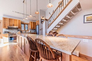 A kitchen with wooden cabinets, a large island with bar stools, a stainless steel refrigerator, and a staircase with wooden railings.