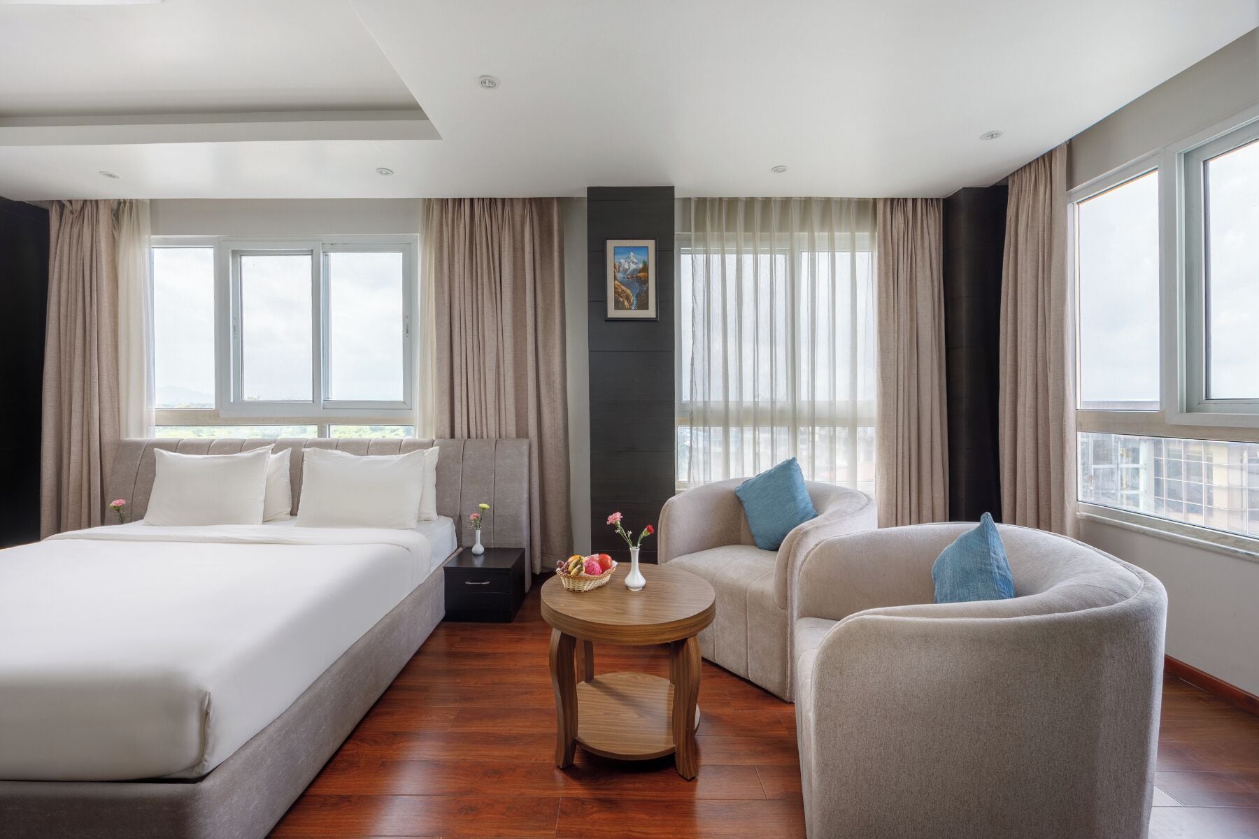 Habitación de hotel con una cama grande, un sofá, una mesita redonda y vistas al exterior a través de amplias ventanas.