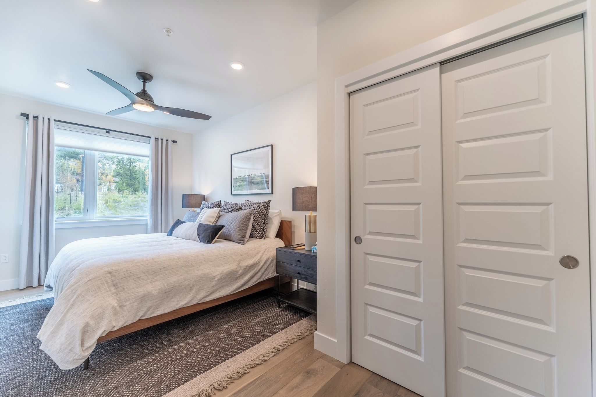 A bedroom with a bed, a ceiling fan, a window with curtains, a nightstand, and a door.
