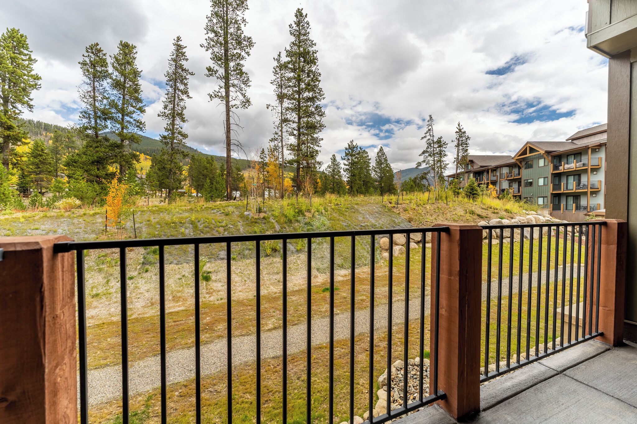 A wooden balcony with a metal railing overlooking a grassy area with trees and a building with balconies.