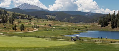 Um campo de golfe com um lago, montanhas e céu claro.
