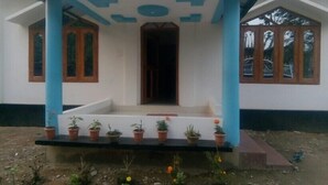 A building with a porch supported by blue columns, a central door, and potted plants on the steps. - Homocation Panchami Homestay (Bokakhat)
