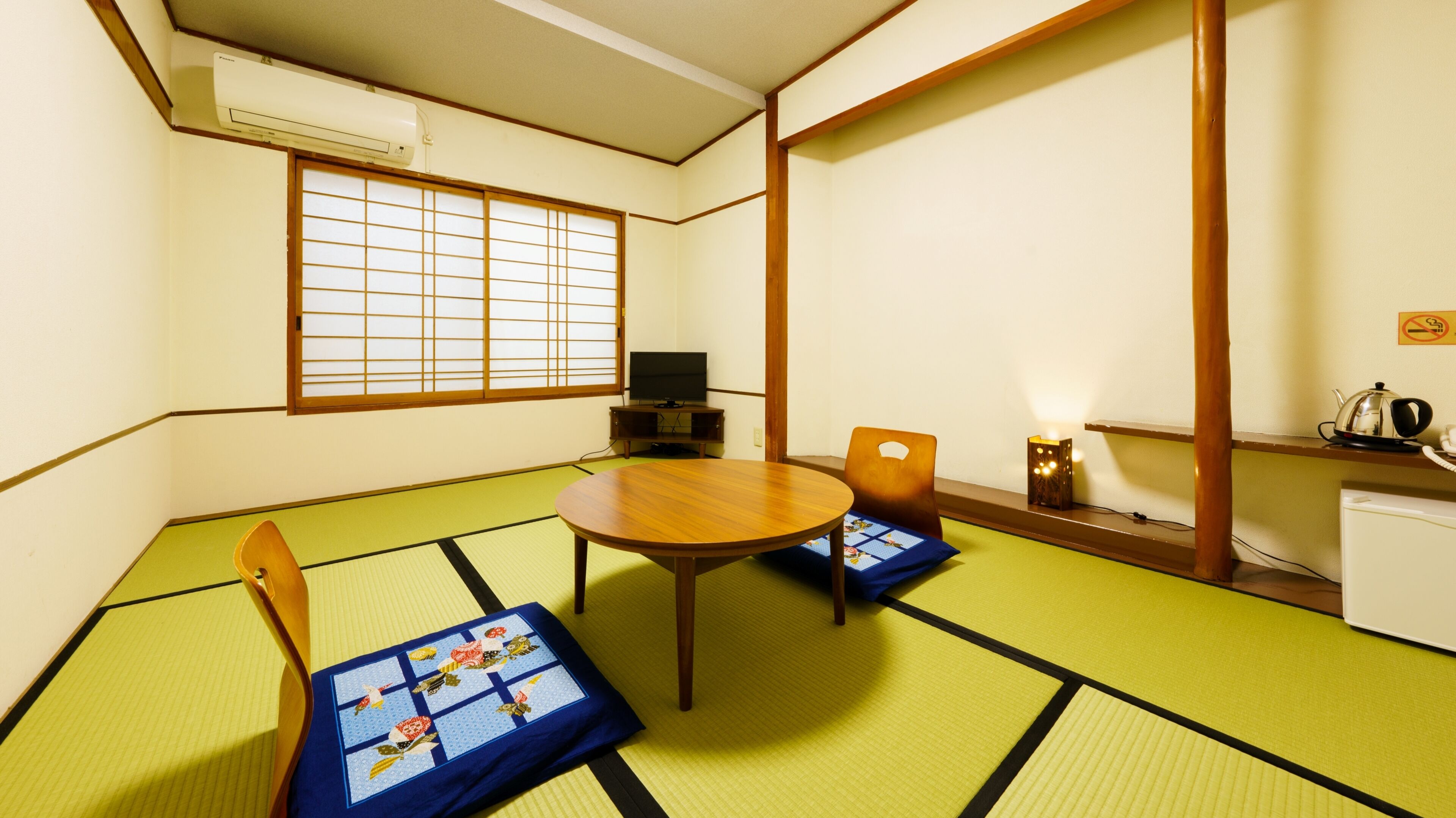 Een kamer met tatami matten, een rond tafel, twee stoelen, een televisie, een waterkoker en een klein kastje.