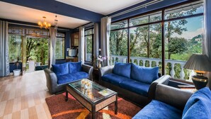 A spacious living room with a large window, blue sofas, a glass coffee table, and a chandelier. - The Junglee Bakri (Mussoorie)