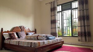 A bedroom with a wooden bed, patterned bedding, a red carpet, and a window with curtains. - Nice Rooms, Unbeatable Prices (Ruhengeri)
