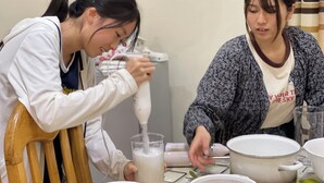 Two individuals at a table, one using a kitchen appliance to prepare a mixture, and the other handling a pot. - Nice Rooms, Unbeatable Prices (Ruhengeri)