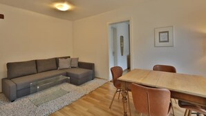 Uma sala de estar com um sofá modular cinza, uma mesa de centro de vidro, uma mesa de jantar de madeira com quatro cadeiras e uma porta que dá para outro cômodo.