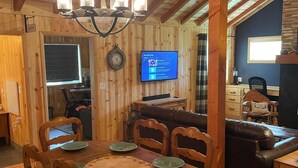 A wooden interior with a dining table set for four, a television displaying a music playlist, and a cozy seating area with a leather couch. - 3 Cabins, 2 Acres, Private Pond, Walk to Restaurants, 4 Separate Sleeping Units (Greer)
