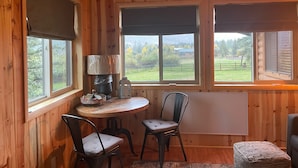 A wooden interior with a round table, two chairs, a lamp, and a view of a green landscape through large windows. - 3 Cabins, 2 Acres, Private Pond, Walk to Restaurants, 4 Separate Sleeping Units (Greer)