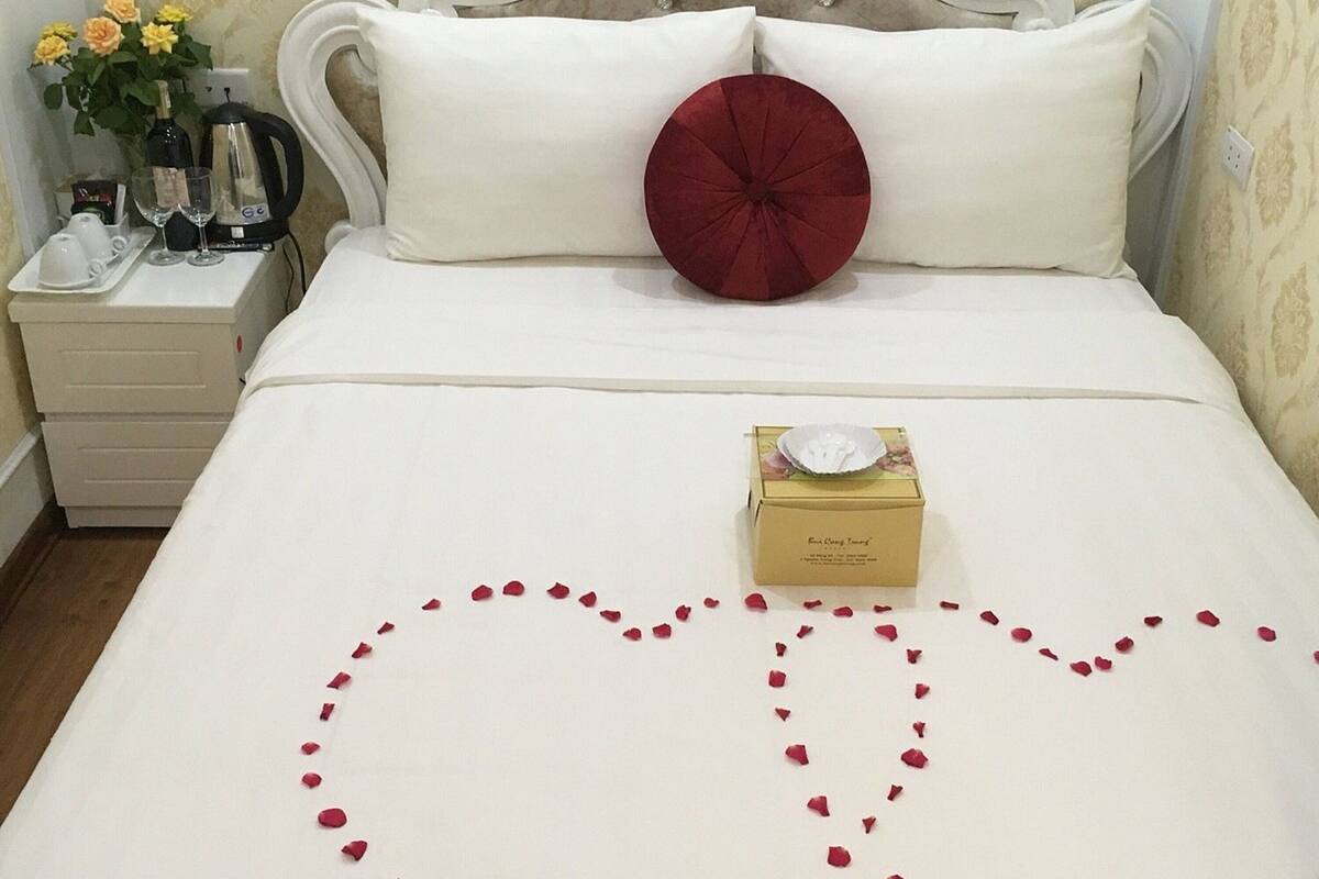 Un lit bien fait, avec du linge de lit blanc, un cœur rouge en guise de décoration posé sur le lit, et une petite boîte ornée de motifs floraux sur le pied de lit.