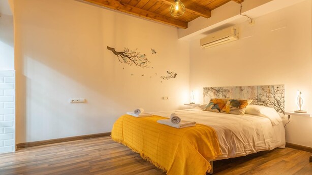 Une chambre avec un plancher en bois, un lit recouvert d’une couverture jaune, un climatiseur fixé au mur et un luminaire au plafond.