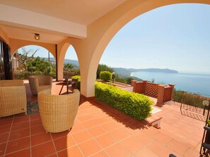 Um terraço com mobiliário de vime, vista para o mar e um jardim paisagístico.
