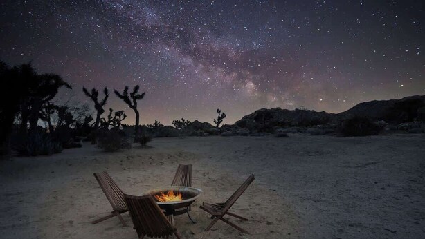 A desert landscape at night with a starry sky, a campfire, and three chairs. - JT Starry Night Getaway Pet-Friendly (Joshua Tree)