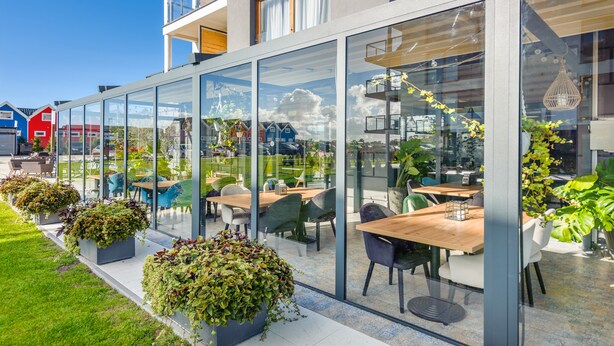 Ein Straßencafé mit Glaswänden, Holztischen und Topfpflanzen.