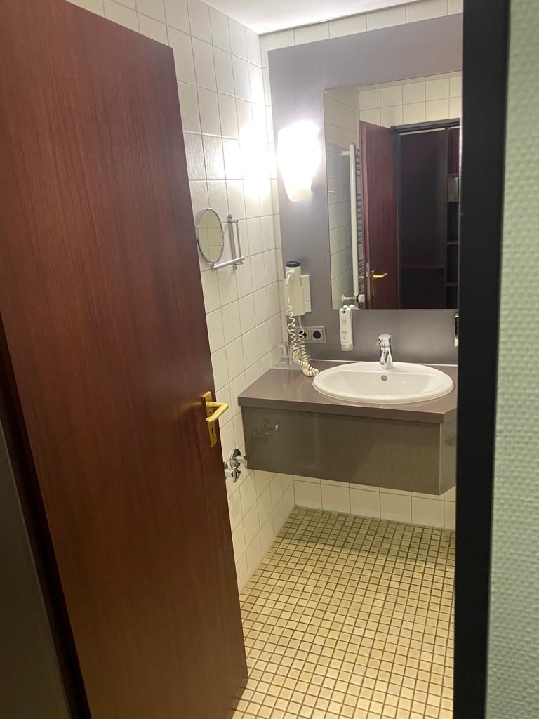 Un baño con puerta de madera, un lavabo blanco, un espejo y suelo de baldosas.