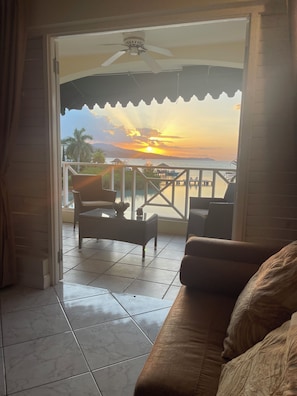 Un balcon avec vue sur un coucher de soleil sur l’eau, un ventilateur de plafond et un coin salon avec un canapé et des fauteuils.