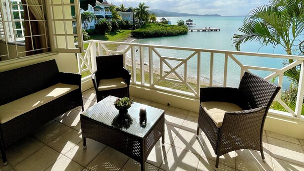 Un balcon doté d’un ventilateur de plafond, d’un canapé, de deux fauteuils en osier et d’une petite table sur laquelle est posée une plante. Le balcon offre une vue sur une étendue d’eau et une île lointaine.