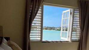 Une chambre avec un grand lit, des oreillers de couleur beige et une vue sur une plage et des maisons, visible par une fenêtre ouverte.