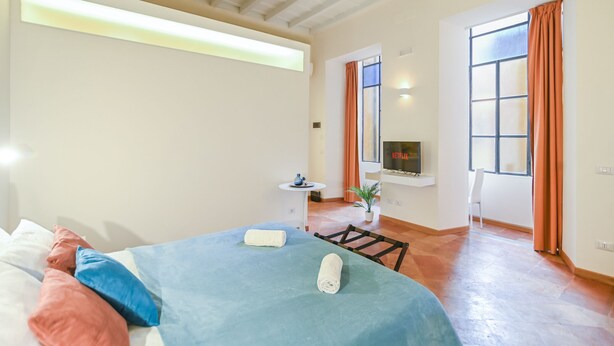 Une chambre à coucher avec un lit, des oreillers, une télévision, une petite table et une fenêtre avec des rideaux.