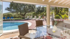 Zona junto a la piscina con sillas de mimbre y una mesa de vidrio, con vistas a una piscina y un jardín ajardinado.
