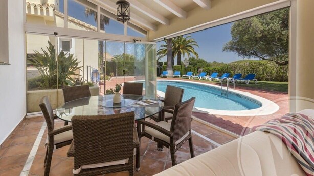 Un patio con paredes de vidrio, una piscina, muebles de mimbre y vistas del paisaje circundante.
