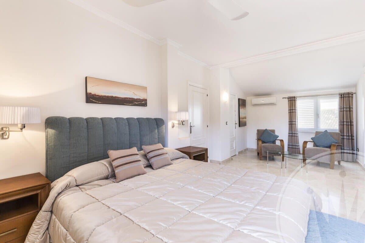 A spacious bedroom with a large bed, a headboard, a bedside table, a nightstand, a chair, a small table, a lamp, a painting, an air conditioning unit, and a window with curtains.