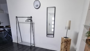 Ein Zimmer mit dunklem Holzboden, einer weißen Wand, einer Uhr, einem Spiegel, einer Holzsäule und einer Lampe.