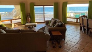 A spacious living room with a large window overlooking the beach, furnished with a sofa, chairs, and a small table. - Casa Semana- Beachfront home (up to 16 guests!) (Playa La Jolla)