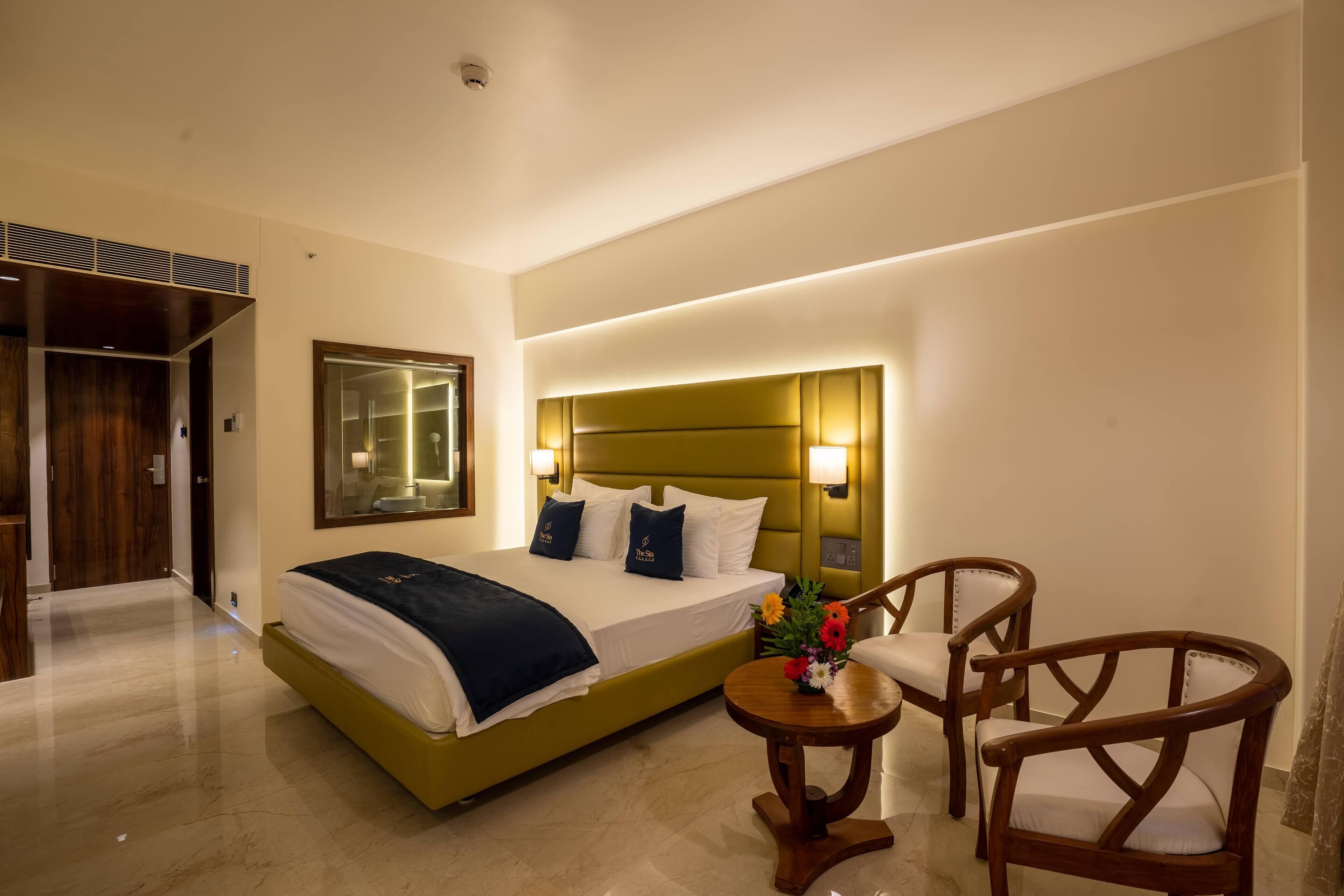 A hotel room with a large bed, bedside lamps, a small round table with a vase of flowers, and two wooden chairs.