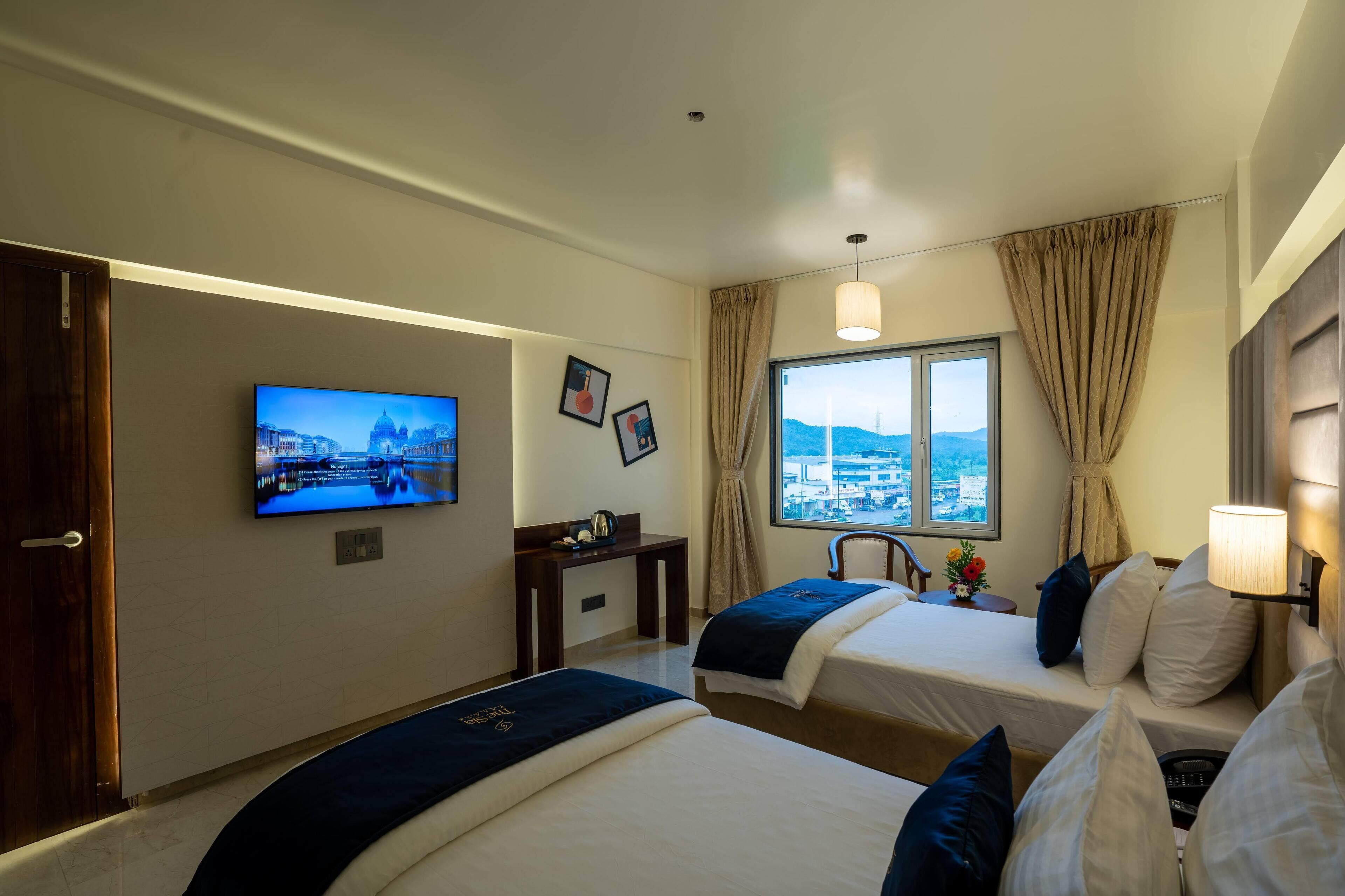 Ein Hotelzimmer mit zwei Betten, einem an der Wand befestigten Fernseher, einem Schreibtisch und einem Fenster mit Blick auf die Stadt.