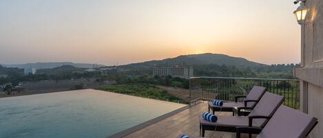 A rooftop pool area with lounge chairs and a view of the sunset over hills.