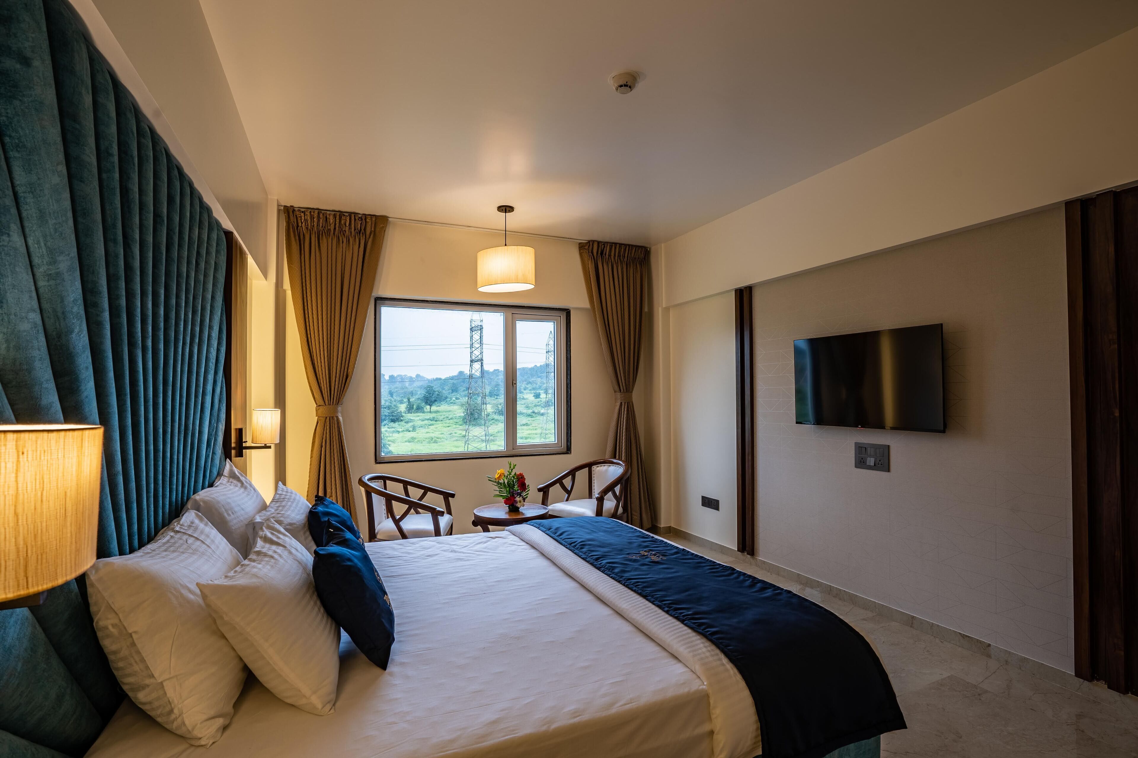 A hotel room with a bed, a TV, a window with curtains, and a view of the outdoors.