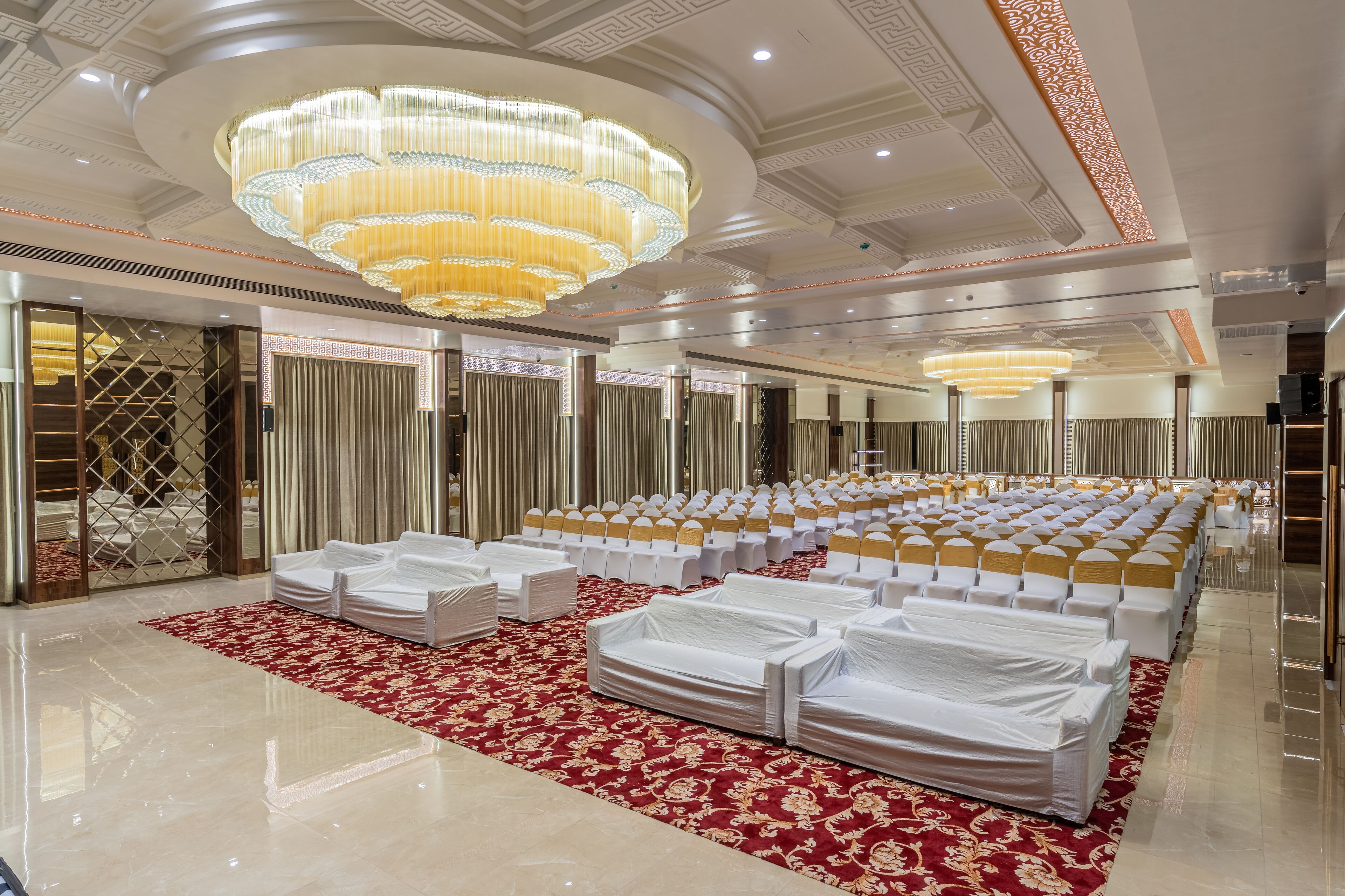 A spacious hall with rows of white chairs and tables, a large chandelier, and a red carpet.