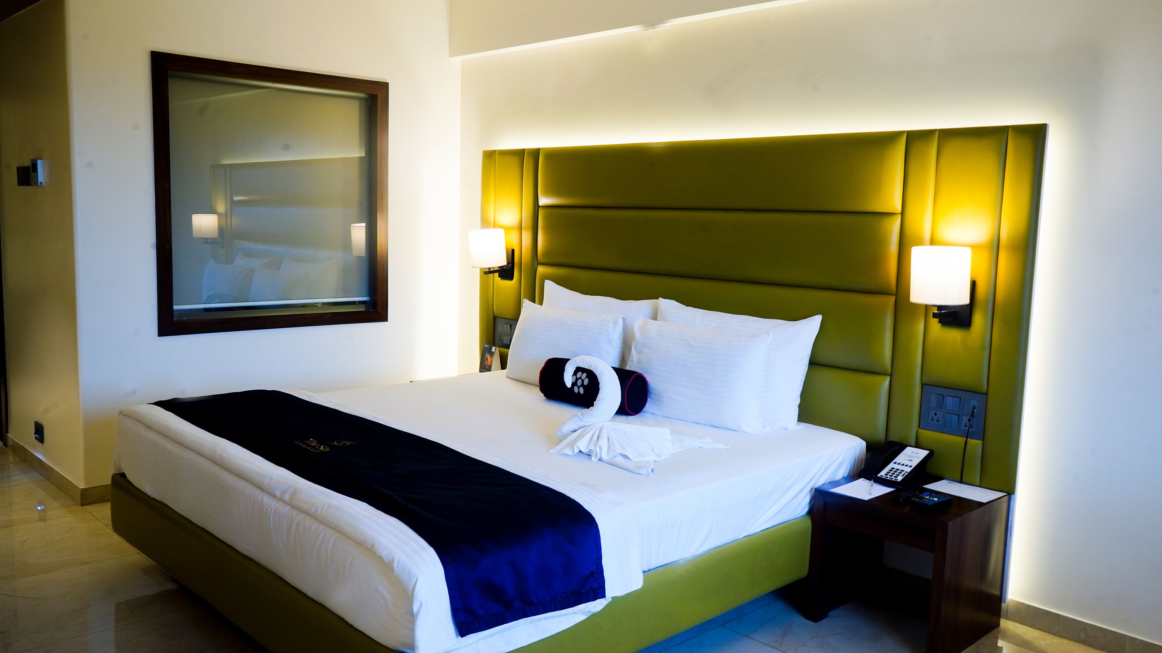 Quarto de hotel com uma cama grande, cabeceira verde, duas luminárias de cabeceira, telefone ao lado da cama e um quadro na parede.