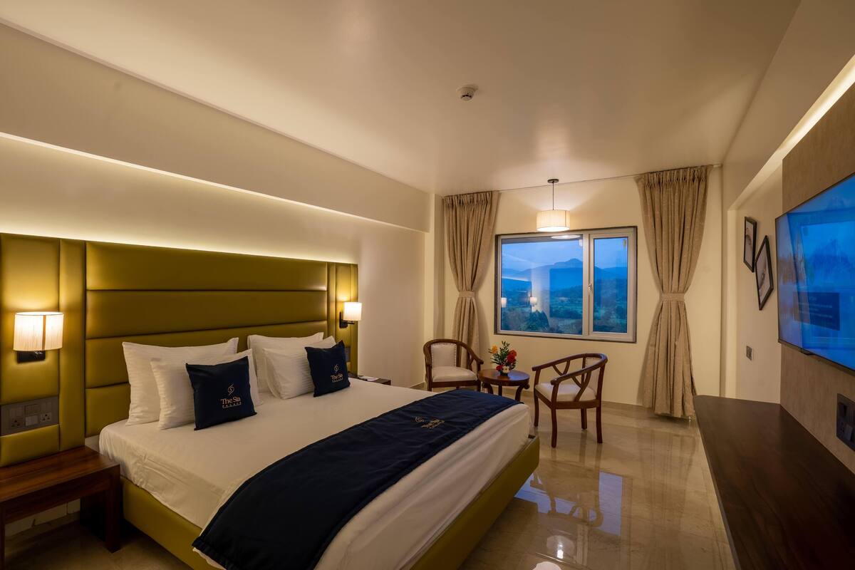 A hotel room with a large bed, bedside lamps, a TV, a desk, and a window with curtains.